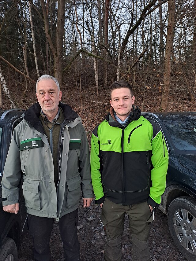 rechts im Bild ein junger Mann in leuchtend grüner Forstjacke und links im Bild ein Herr um 60 mit olivgrüner Forstjacke