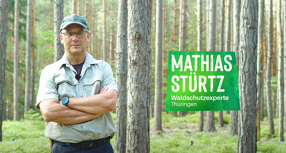 Forstmitarbeiter um die 50 Jahre mit Brille, grauem Hemd und Schirmmütze im Wald neben einem Baumstamm, der Mann hält die Arme verschränkt.