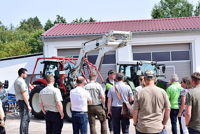 Eine Gruppe von Männern und Frauen betrachtet ein einem Traktor ähnliches forstliches Fahrzeug, währen ein Mann dazu spricht.