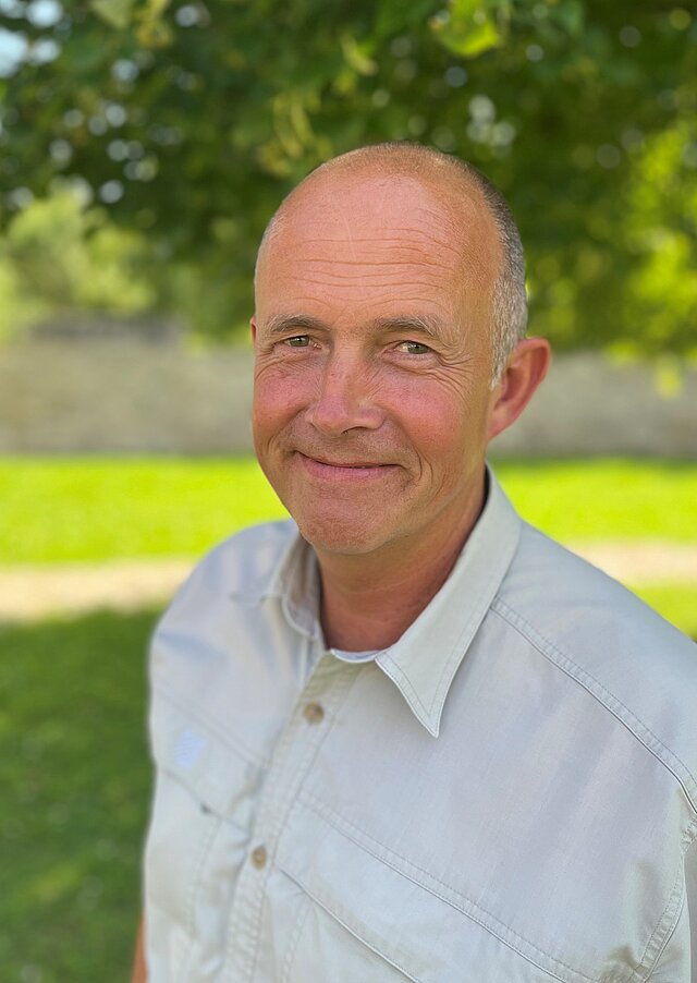 Ein freundlich lächelnder Mann mittleren Alters in einr parkähnlichen, grünen Landschaft.