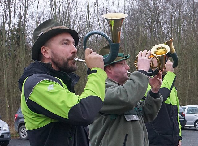 Ein Jäger in neongrüner Jacke mit schwarzen Applikationen und Filzhut bläst ein Jagdhorn